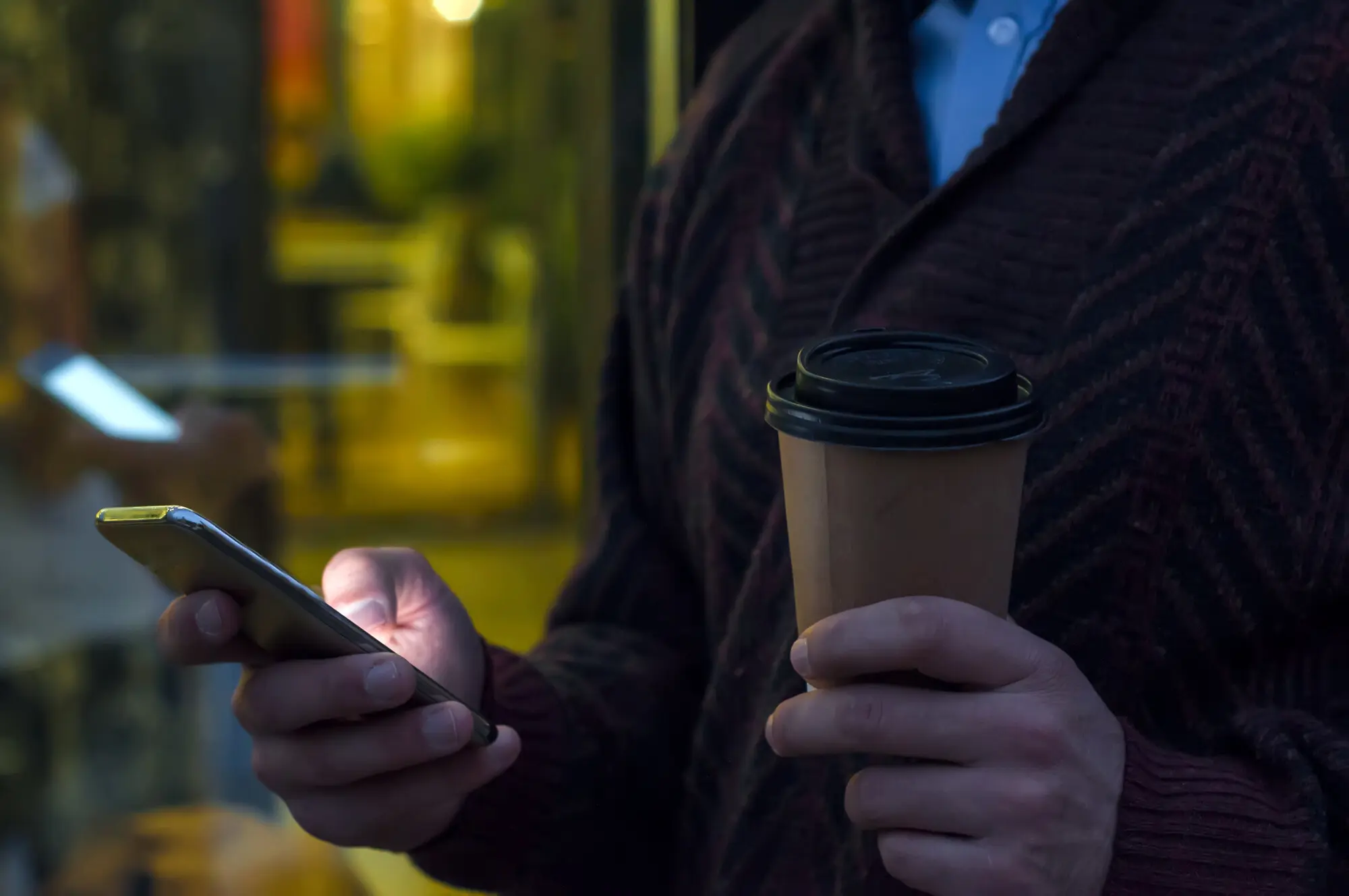 Nahaufnahme eines Geschäftsmanns, der ein Mobiltelefon benutzt und einen Pappbecher hält. Detailaufnahme der Hand des Geschäftsmanns, die einen Pappbecher hält und ein Smartphone benutzt, während er auf der Straße geht, praxisnah und nachvollziehbar.